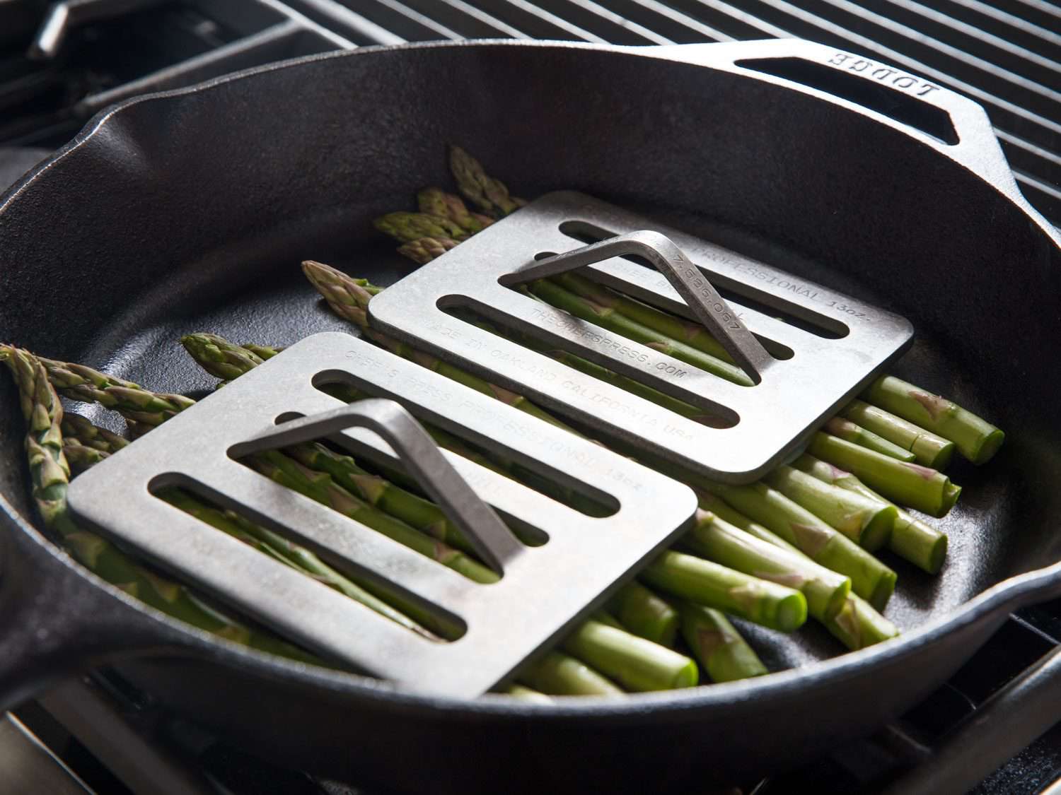 Metal cooking presses on top of asparagus in a cast iron pan.