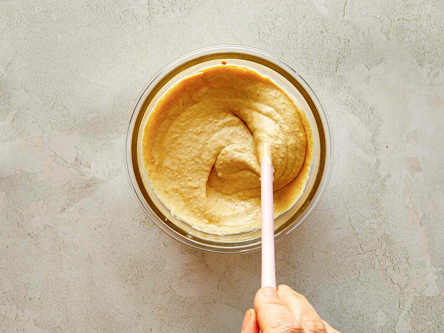 A hand stirring a bowl of creamy mixture with a spatula