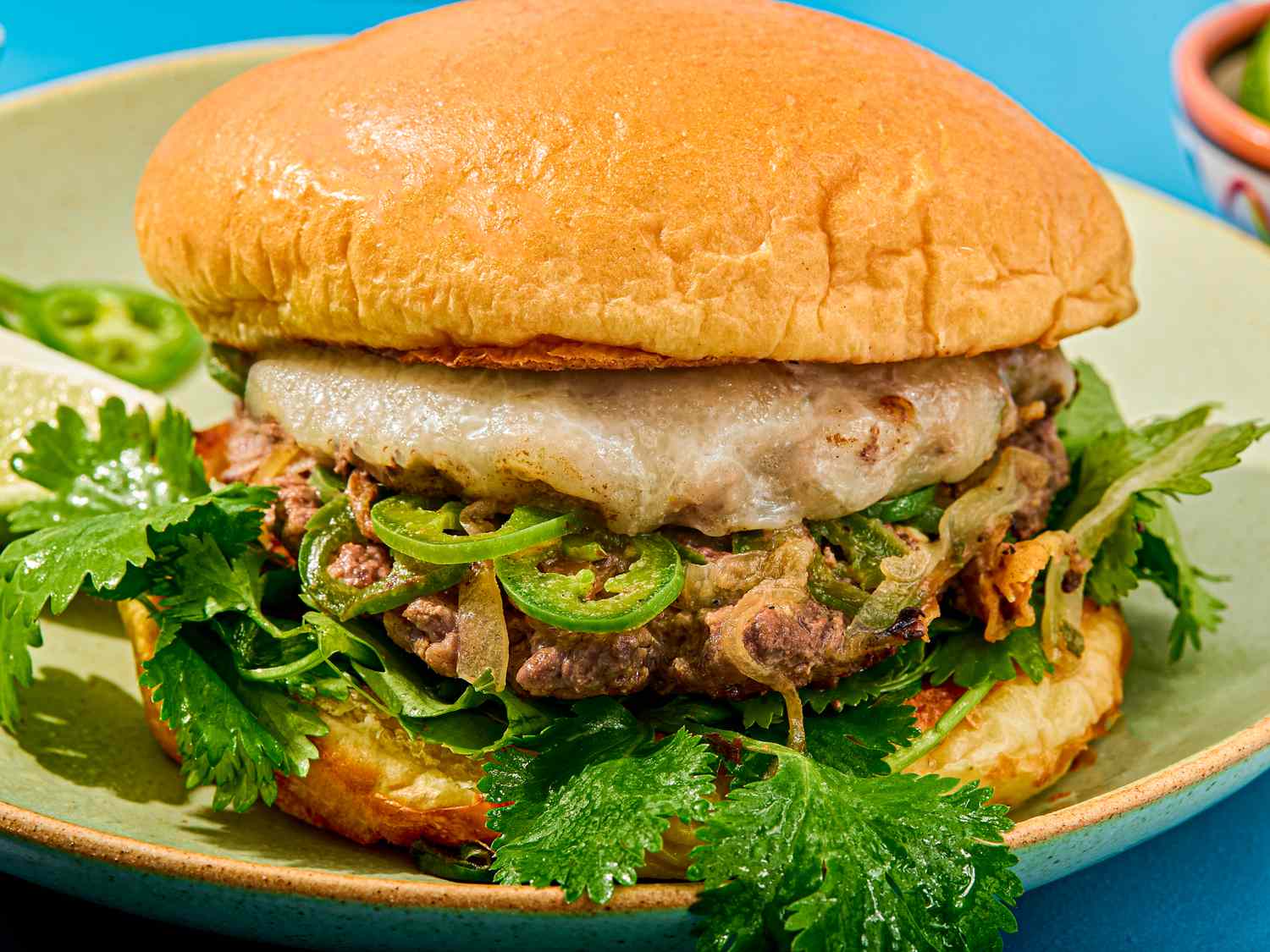 Close-up of a burger with melted cheese, slices of green peppers, cilantro, and a toasted bun on a green plate