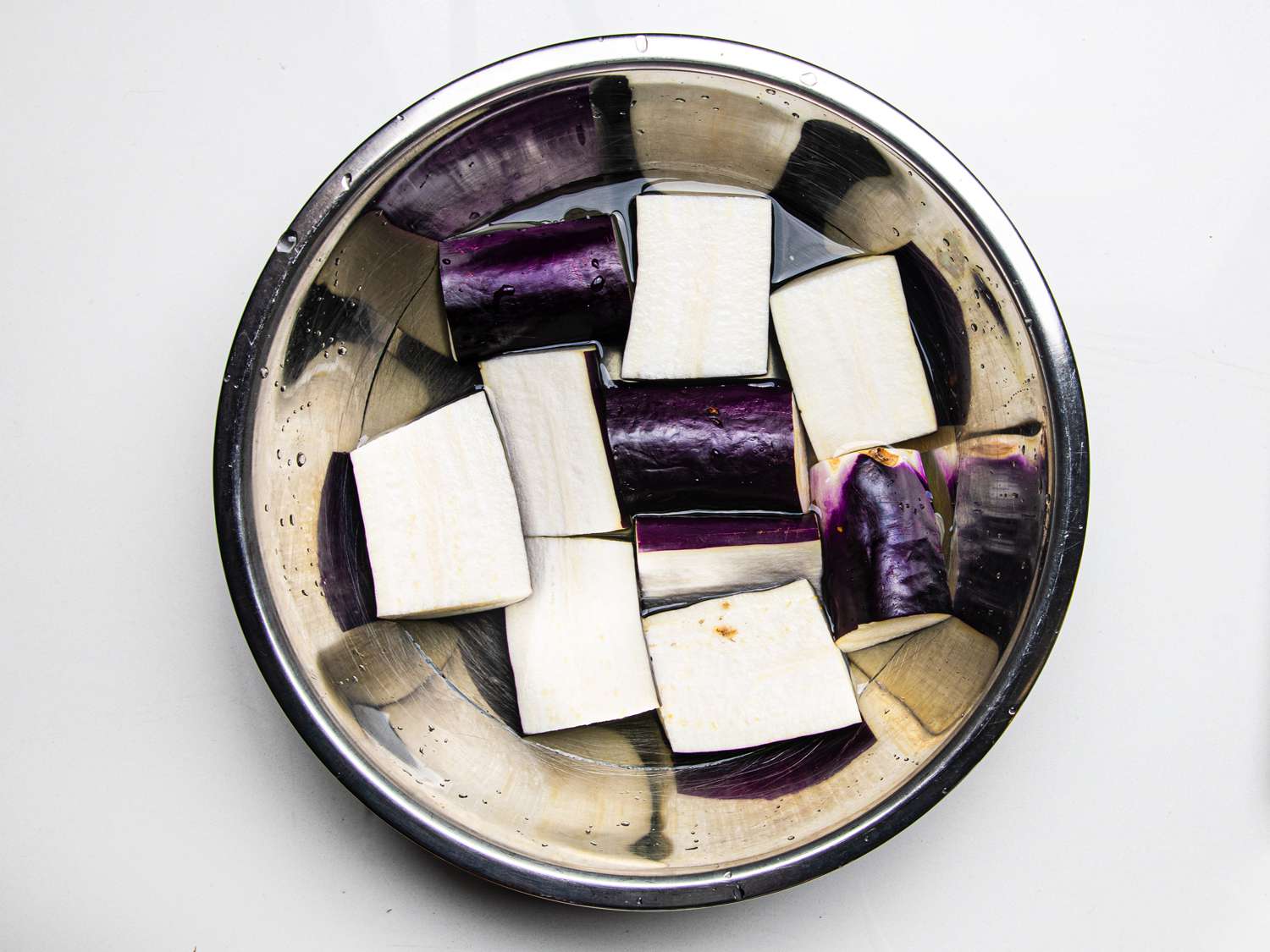 Cut eggplants soaking in a metal bowl on a white counter