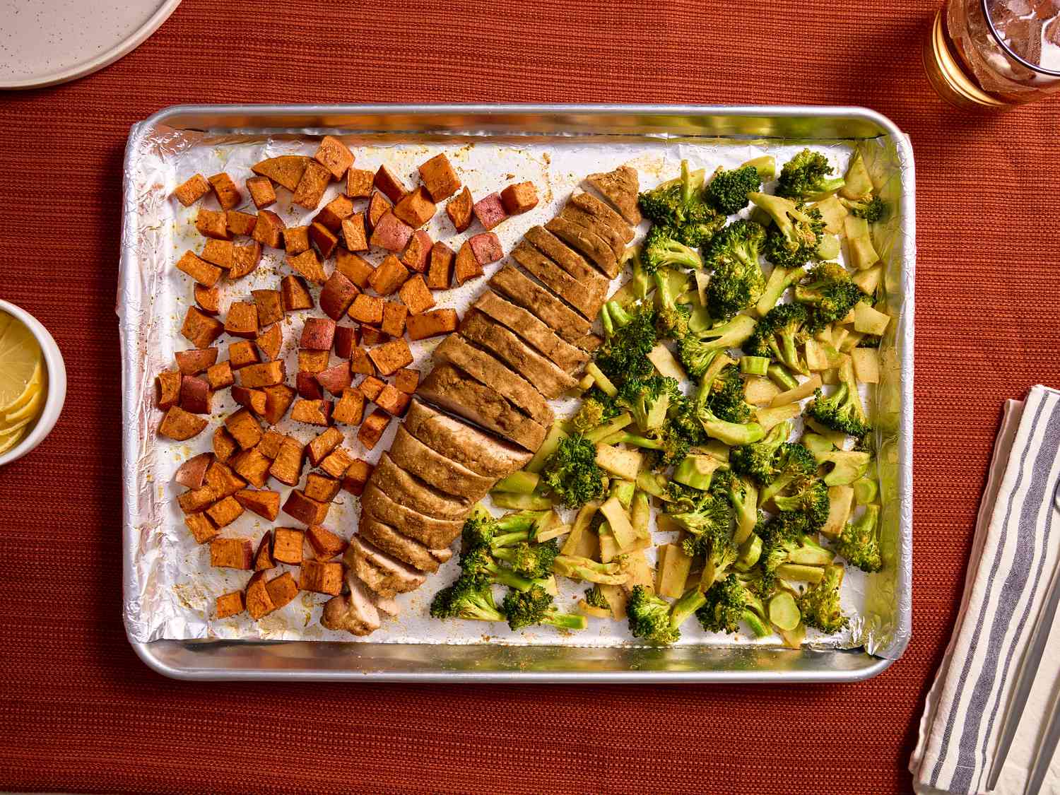 Sheetpan with sloced pork and veggies on red tabletop, with lemon slices plates and napkins on the sides 