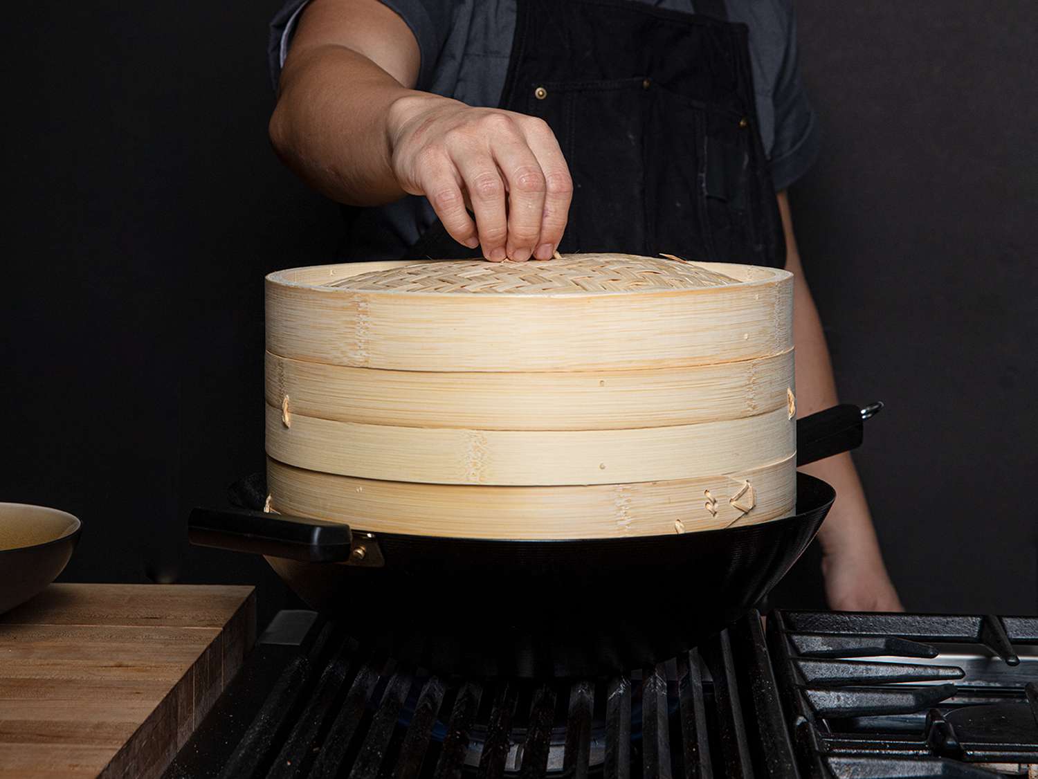 A person about to lift the lid off a two-tier bamboo steamer in a wok