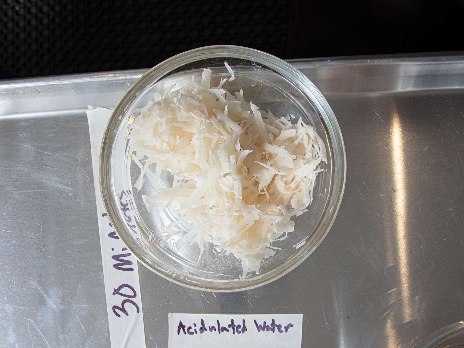 Shredded potato in a glass bowl labeled acidulated water