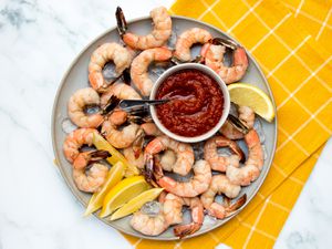 Shrimp cocktail on a ceramic plate with cocktail sauce and sliced lemons, on a stone background with a yellow dish towel.