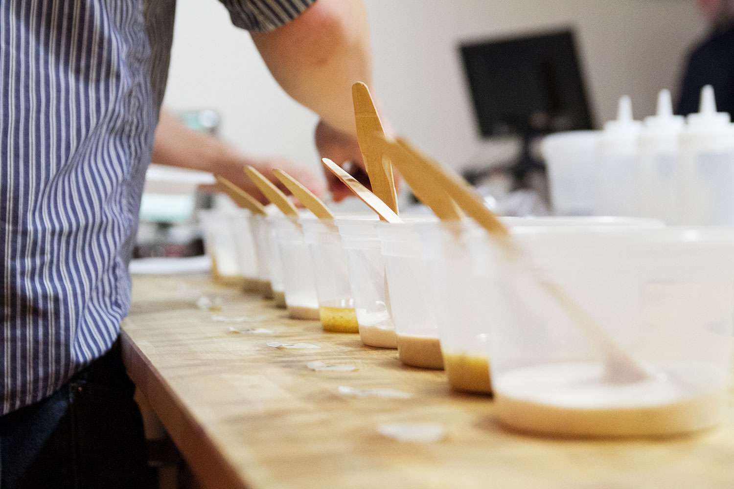 A line of plastic bowls each with a wooden spoon and each containing a different Caesar dressing.