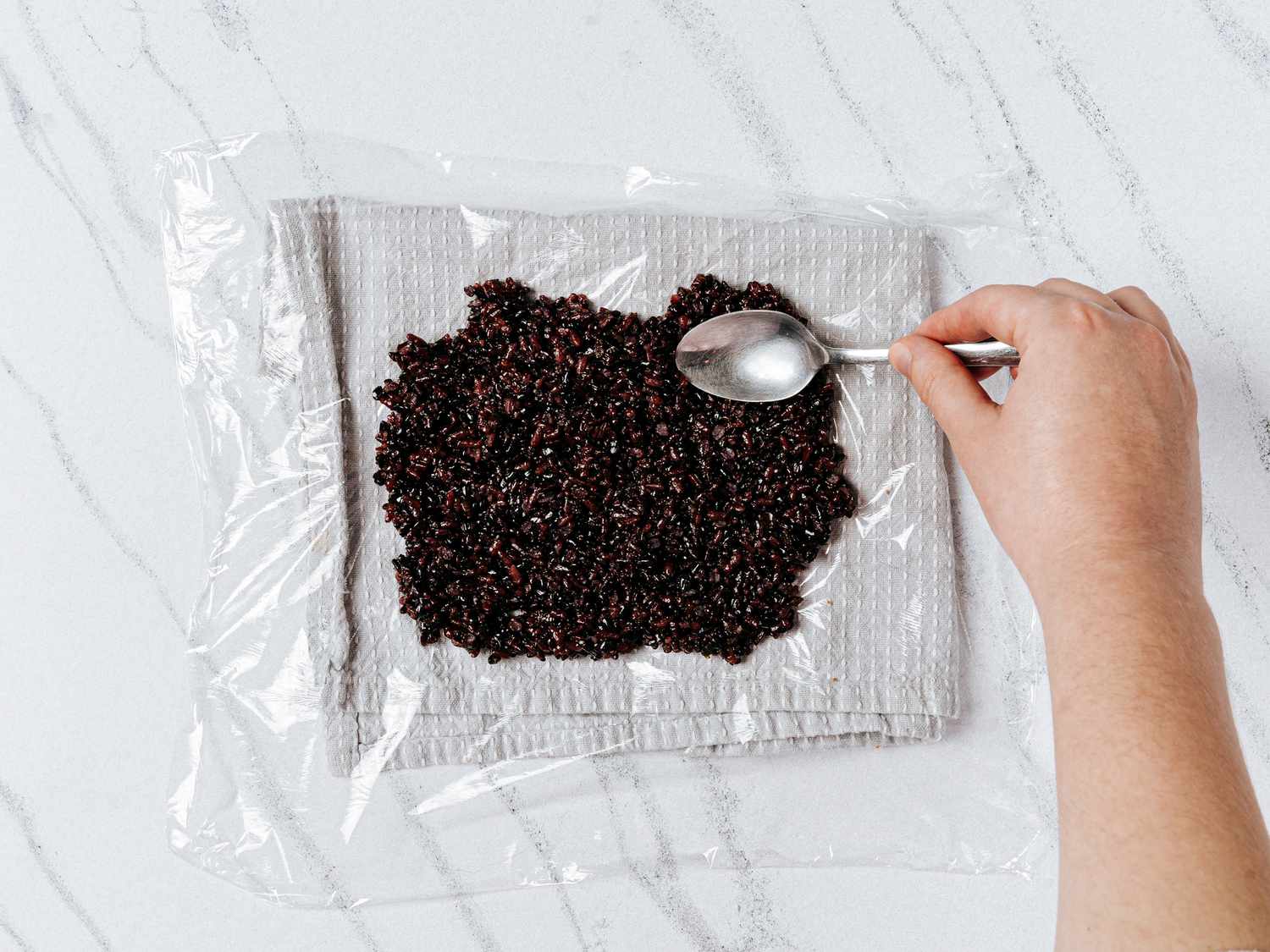 Hand using the back of a spoon to spread rice onto plastic wrap, which is layed out on top of a folded white towel 