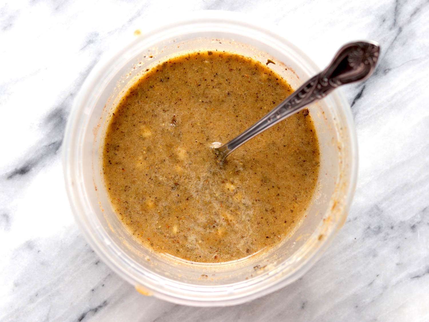 A container of puréed garlic, orange juice, lime juice, cumin, oregano, black pepper, allspice with a spoon in the container.