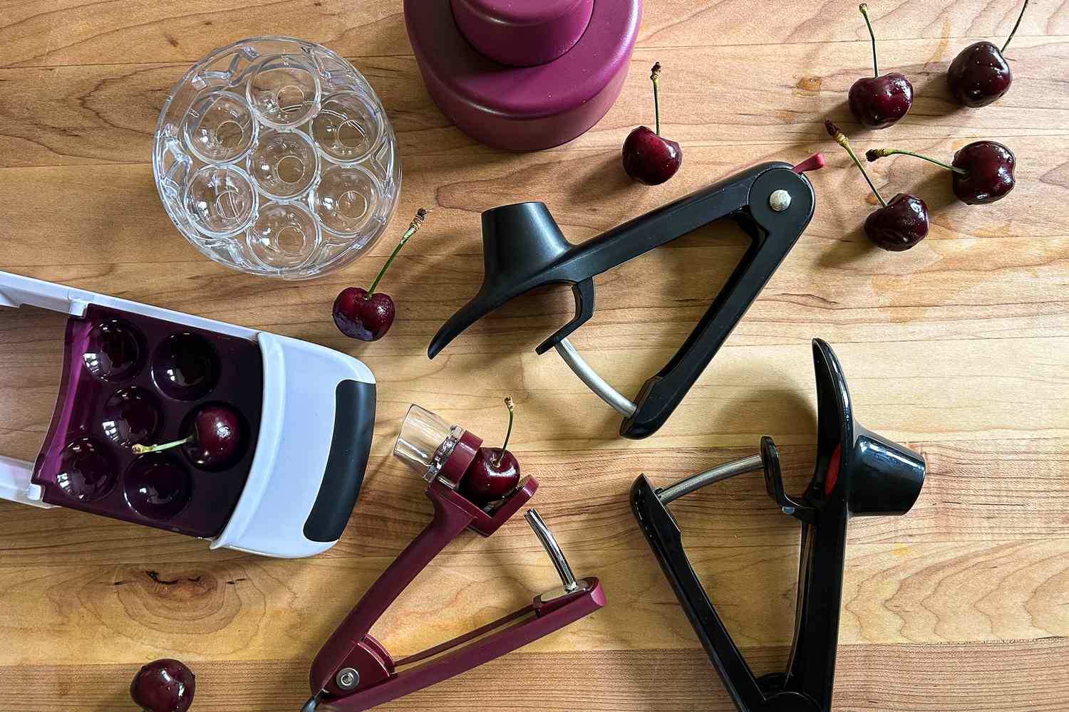Five cherry pitters on a wooden surface with a few cherries strewn around them