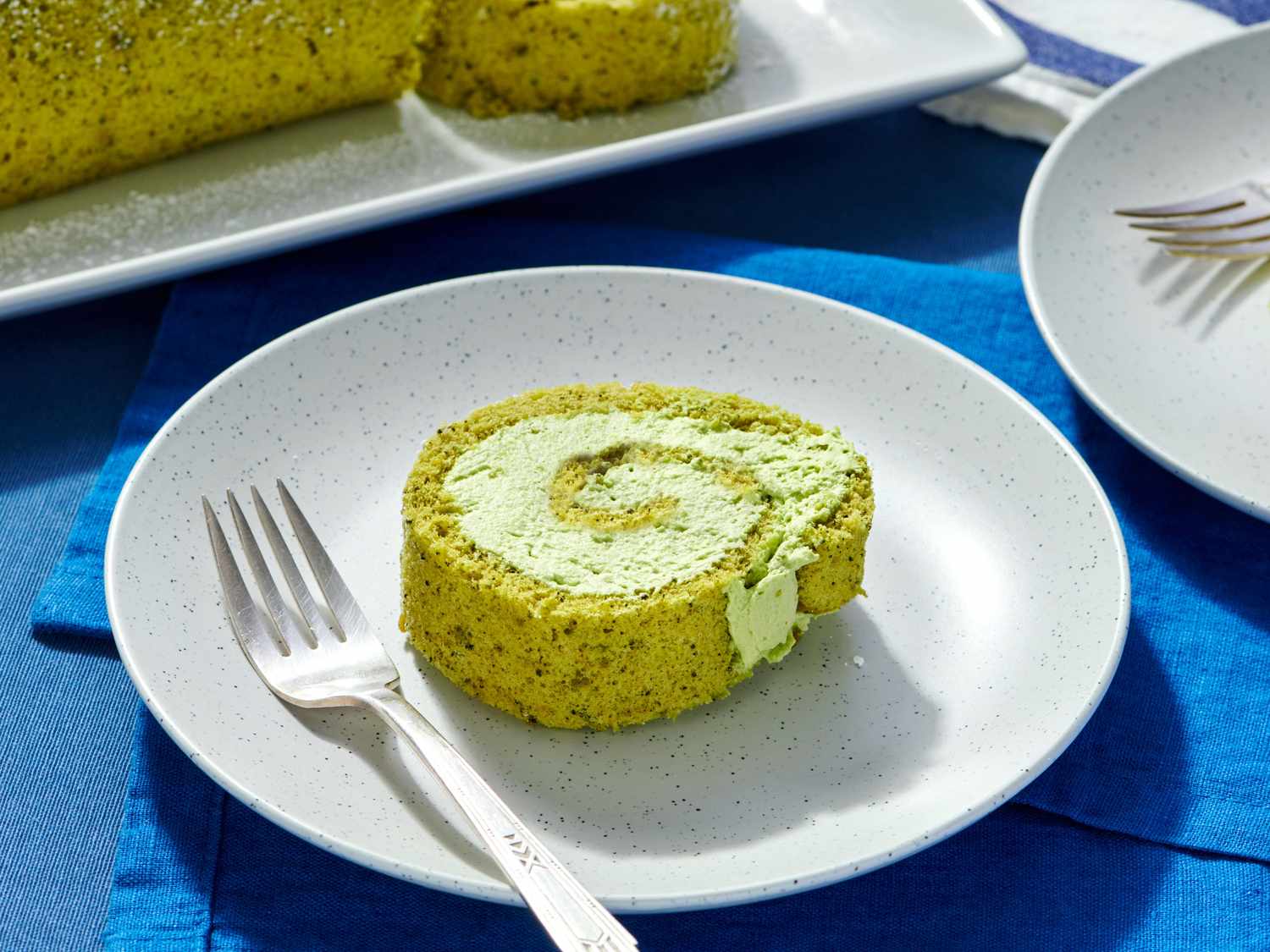 A slice of matcha swiss roll on a plate with a fork with additional rolled cake in the background and blue tablecloth underneath