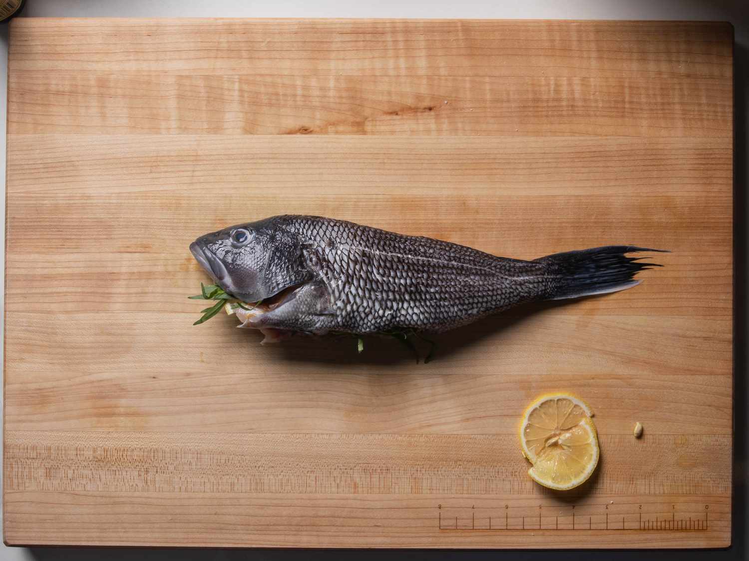 A whole black bass on a cutting board; it's been stuffed with fresh tarragon and lemon slices.