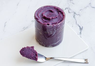 A glass jar of ube halaya placed on a white marble surface. A morsel of the stiff jam is perched on a spoon in front of the jar.