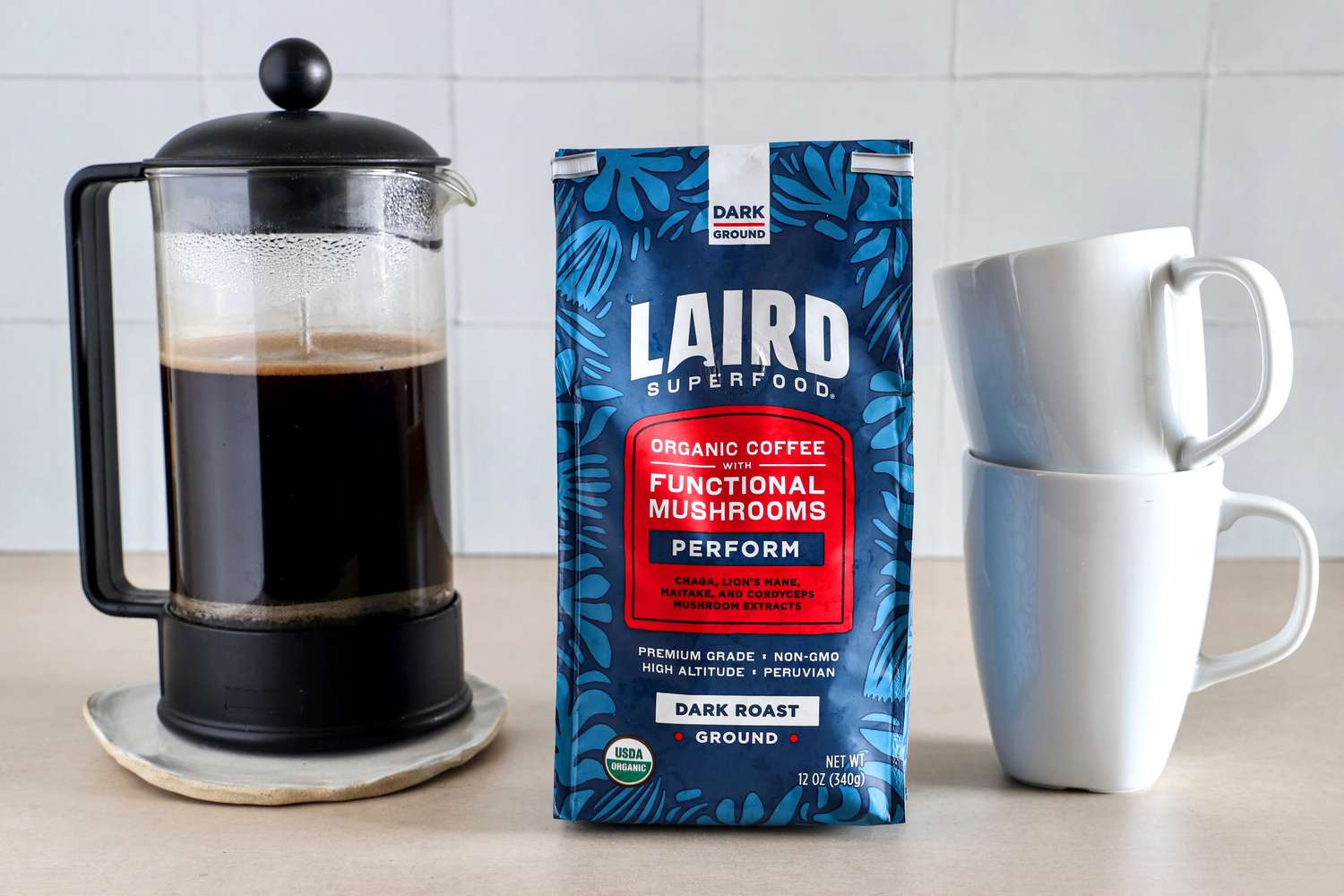 A French press, a package of Laird Superfood coffee, and stacked white mugs on a countertop