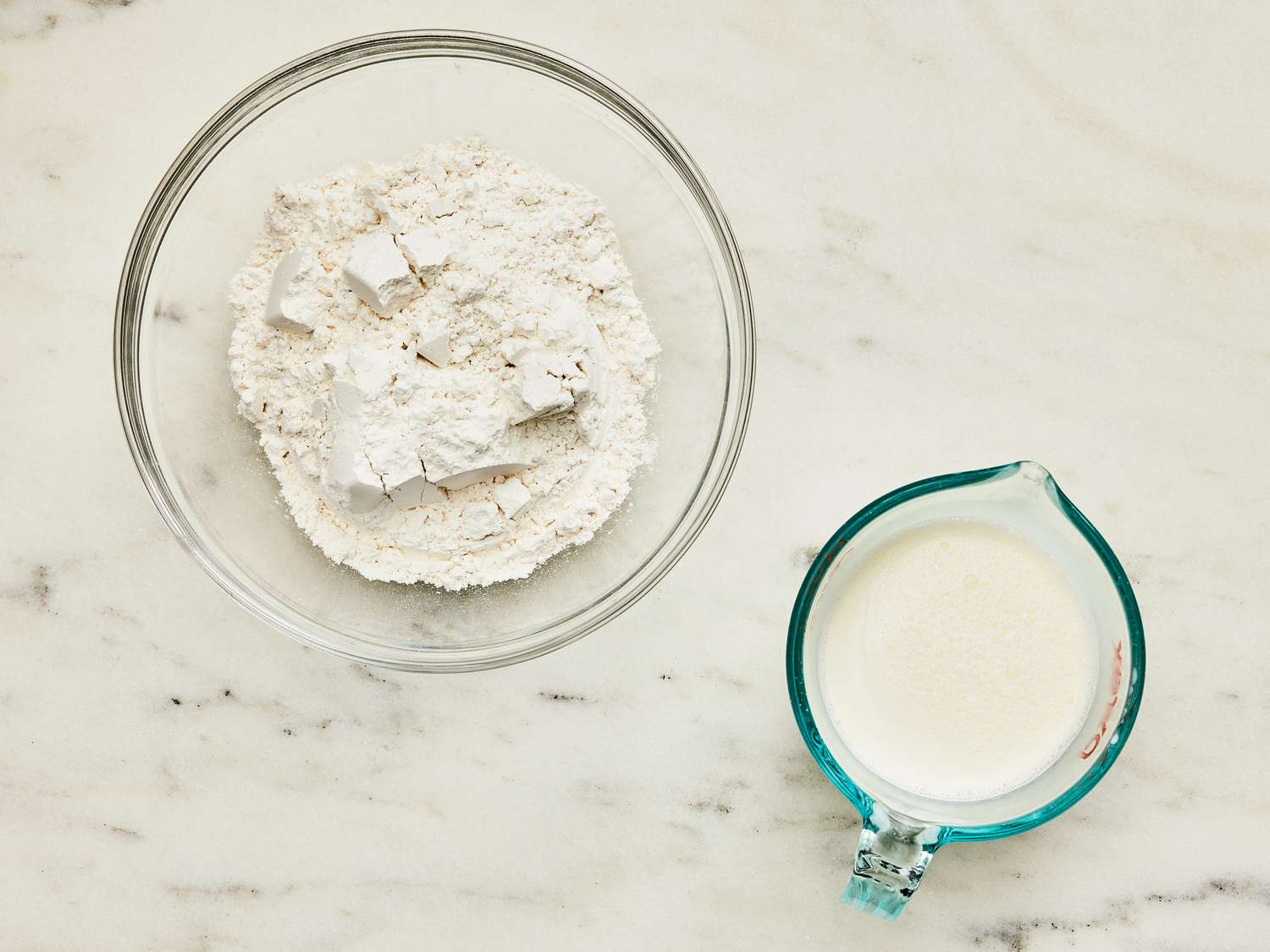 Overhead angle of flour in glass bowl and milk in glass measuring cup on a marble tapletop