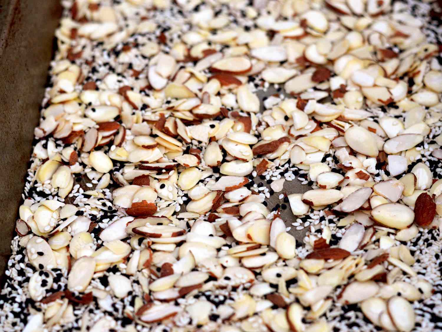 Sliced almonds and white and black sesame seeds cover the bottom of a rimmed baking sheet.