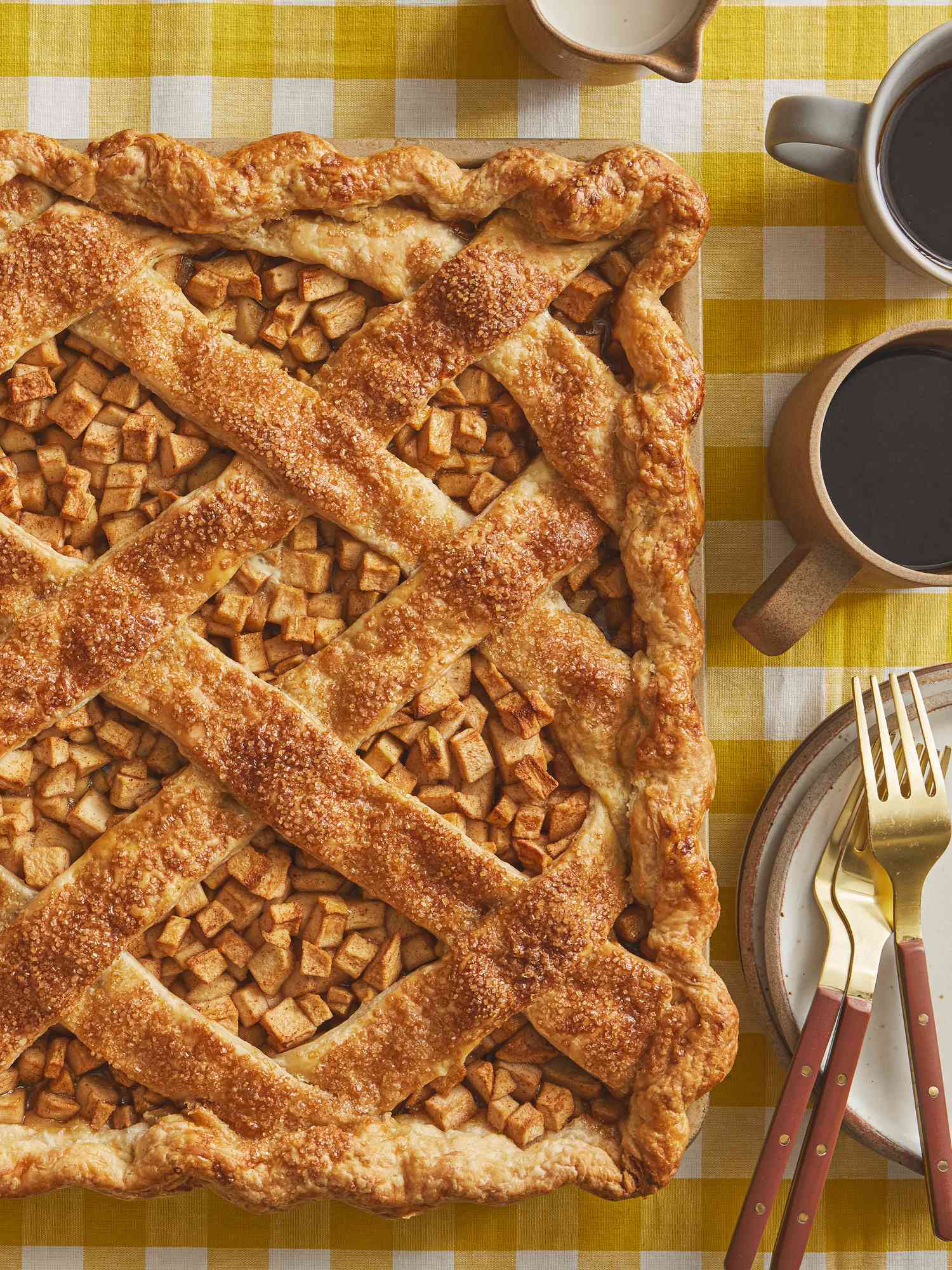 Whole Apple Slab Pie being placed on a plate. Tray is on a yellow gingham print tablecloth, with two cups of coffee, plates and gold forks 