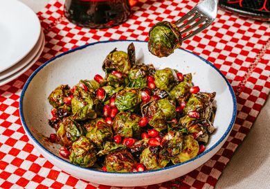 A bowl of airfried Brussels sprouts garnished with pomegranate seeds with a fork picking up a piece placed on a checkered tablecloth