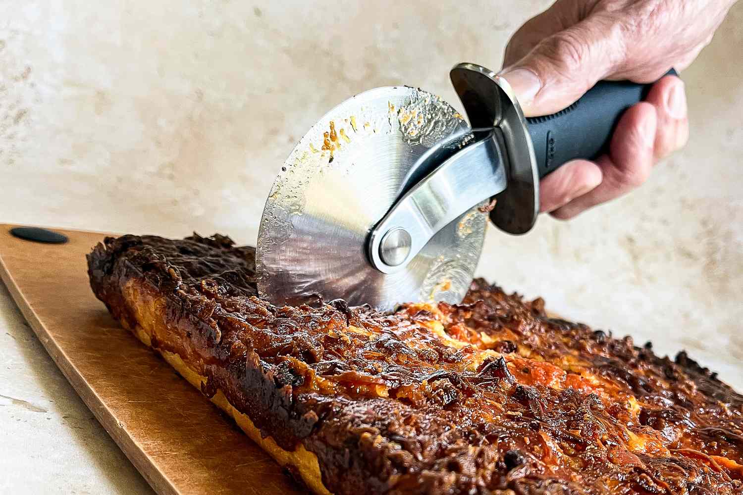 A hand using an OXO pizza cutter to slice baked food on a cutting board