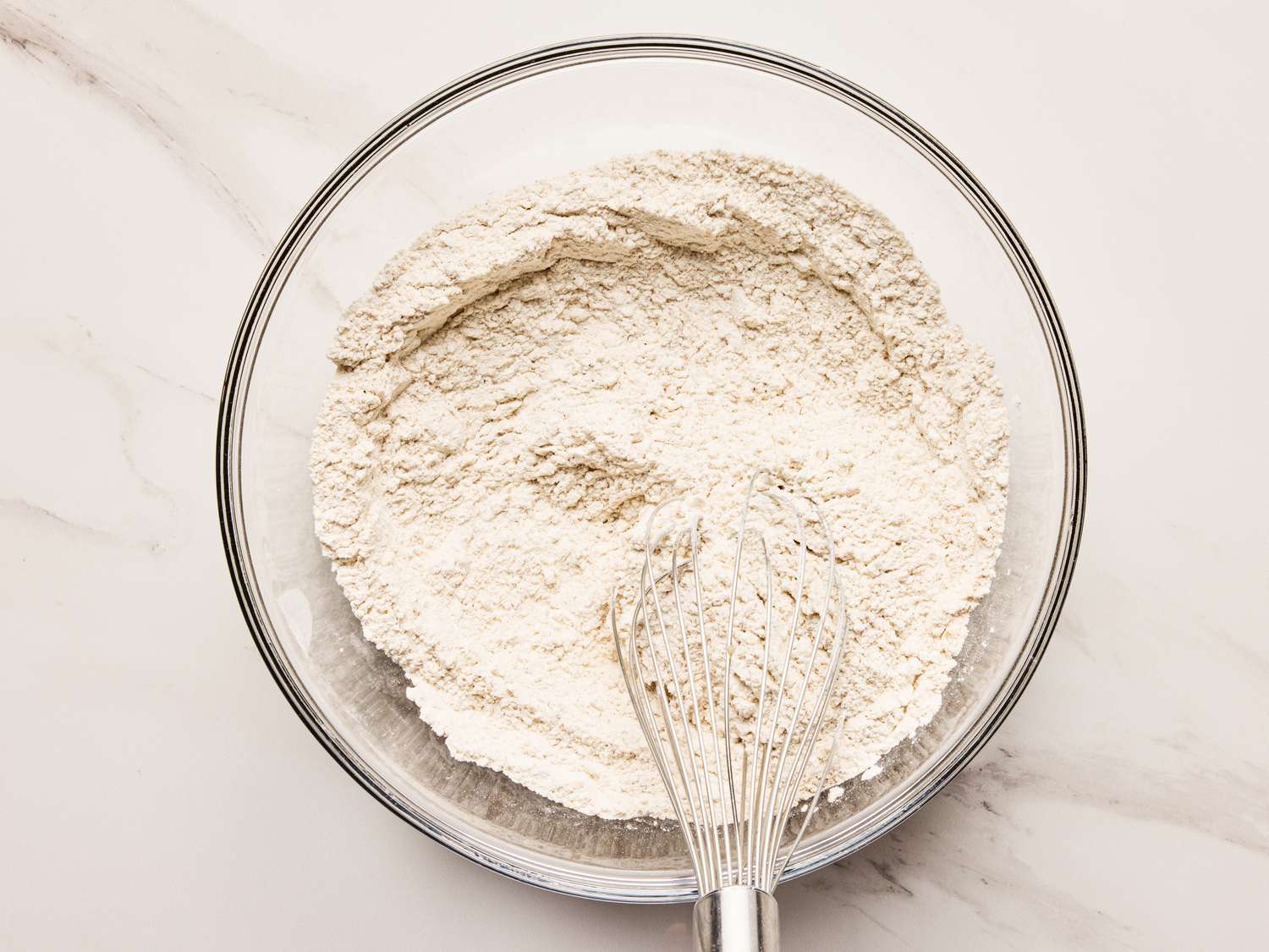 A whisk placed in a bowl of flour on a marble countertop