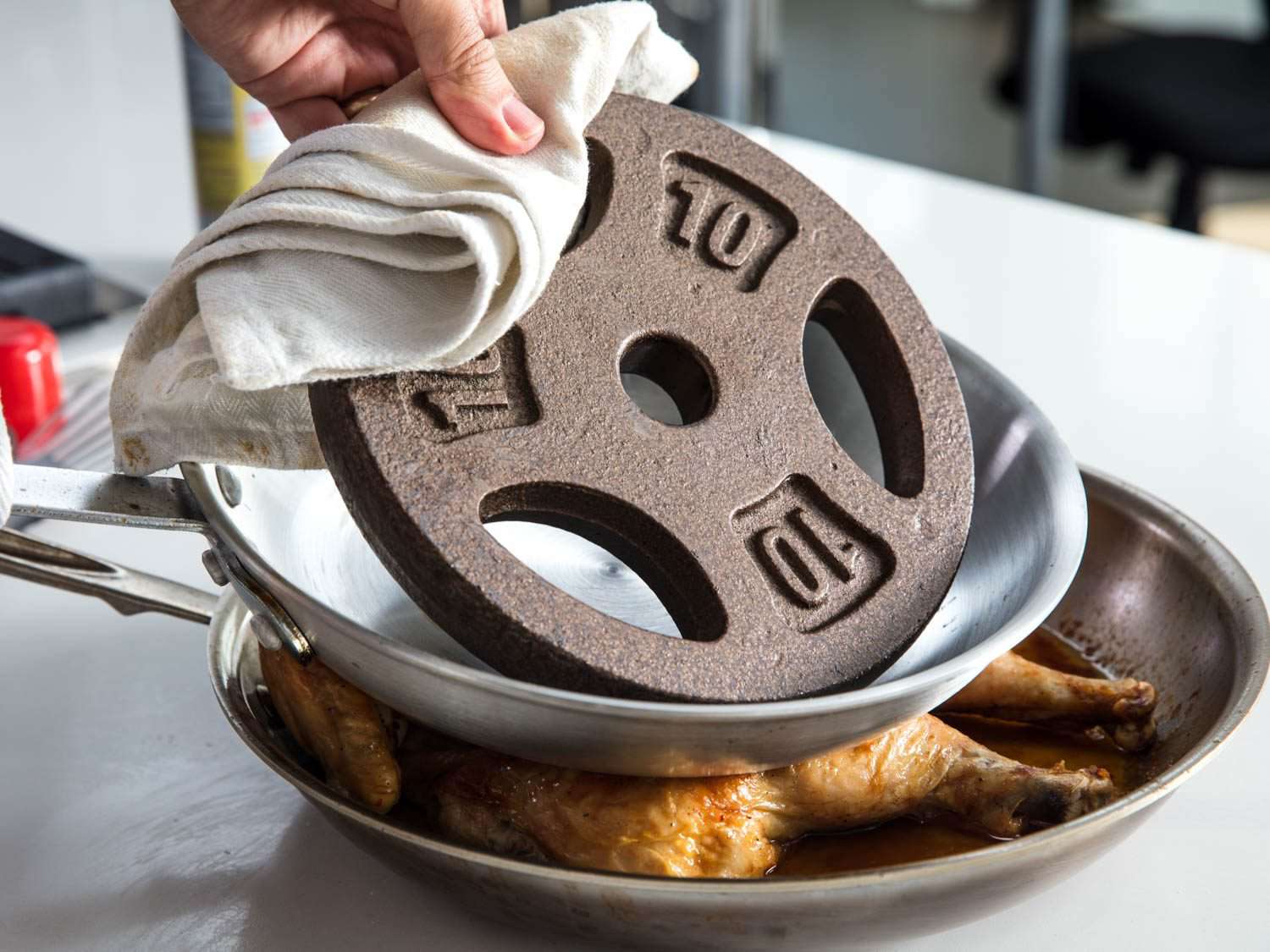 A hand lifting a barbell weight plate from a metal skillet on top of a spatchcocked roast chicken in another skillet.