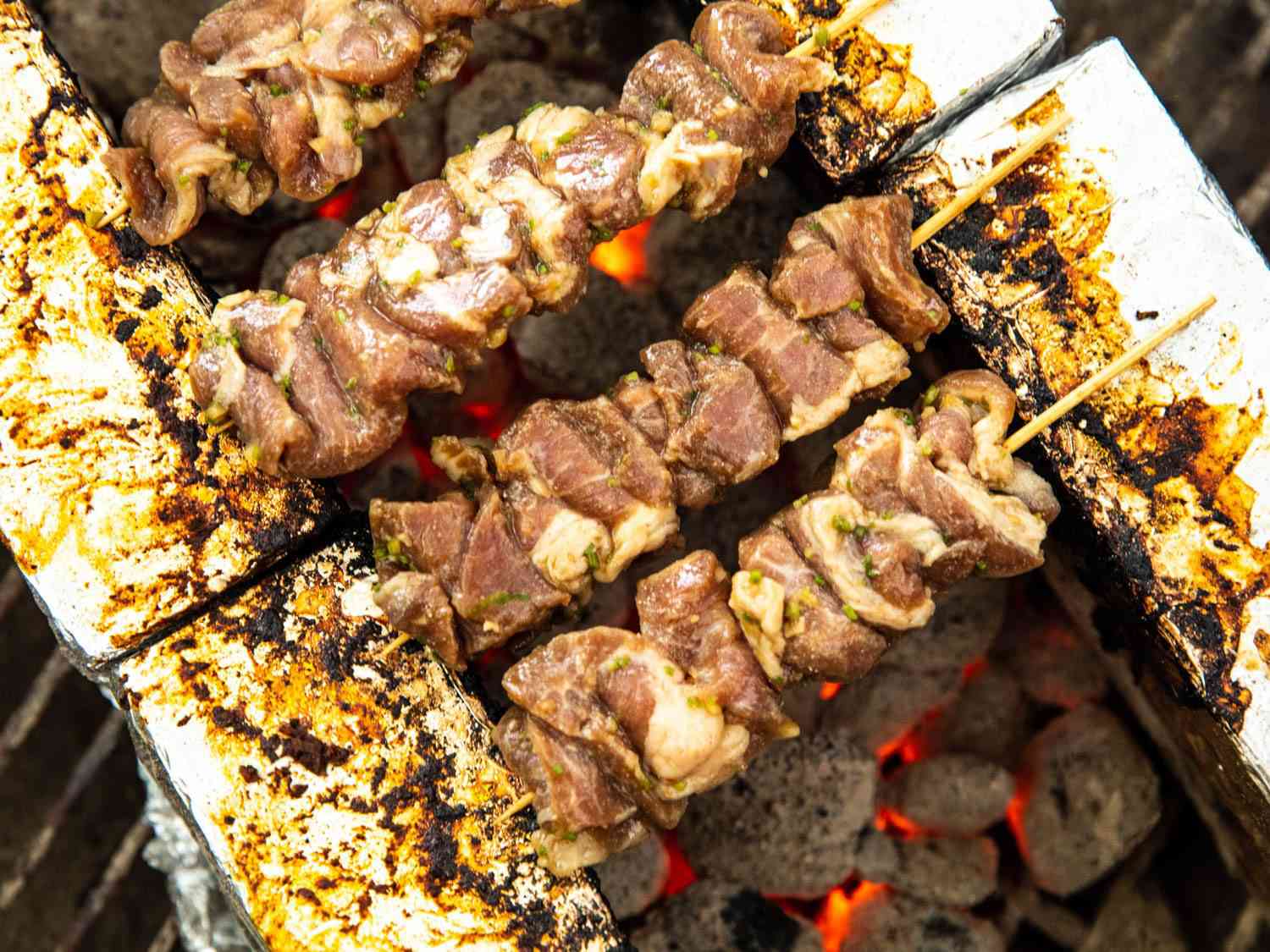 Four partially cooked pork Thai skewers on a brick stand over coals.