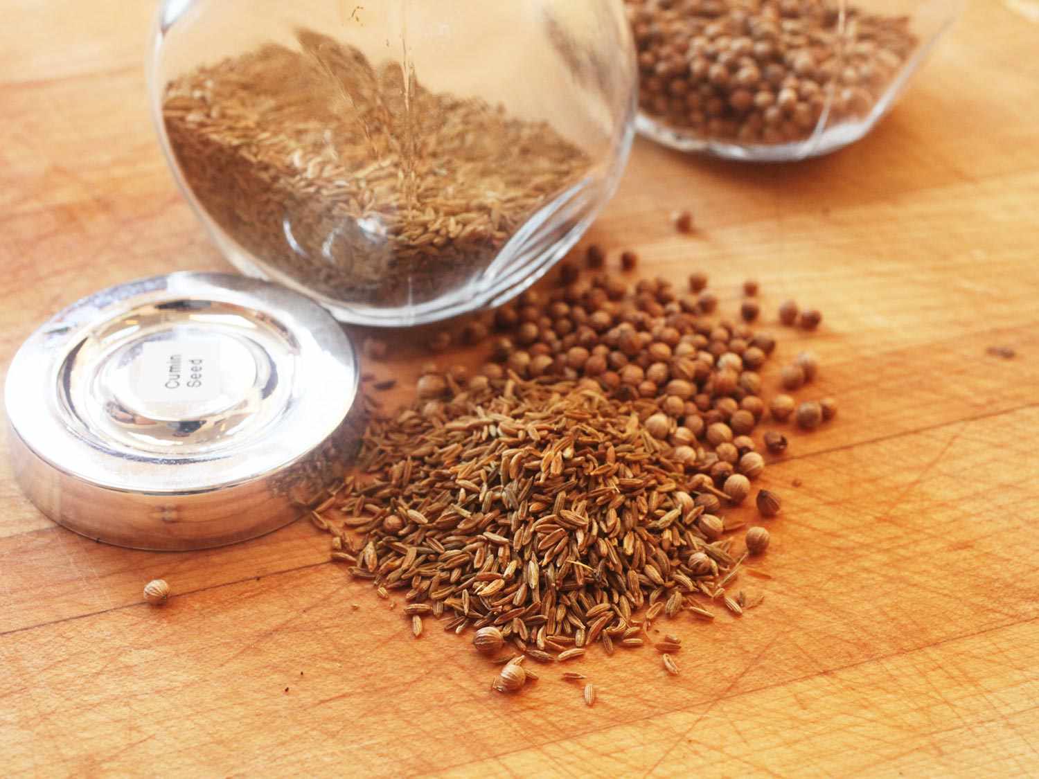 Whole cumin and coriander seeds on a wooden cutting board with their jars in the background.