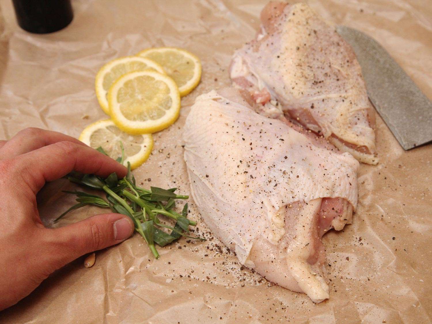 Two chicken breast halves next to lemon slices and chopped herbs and a hand.