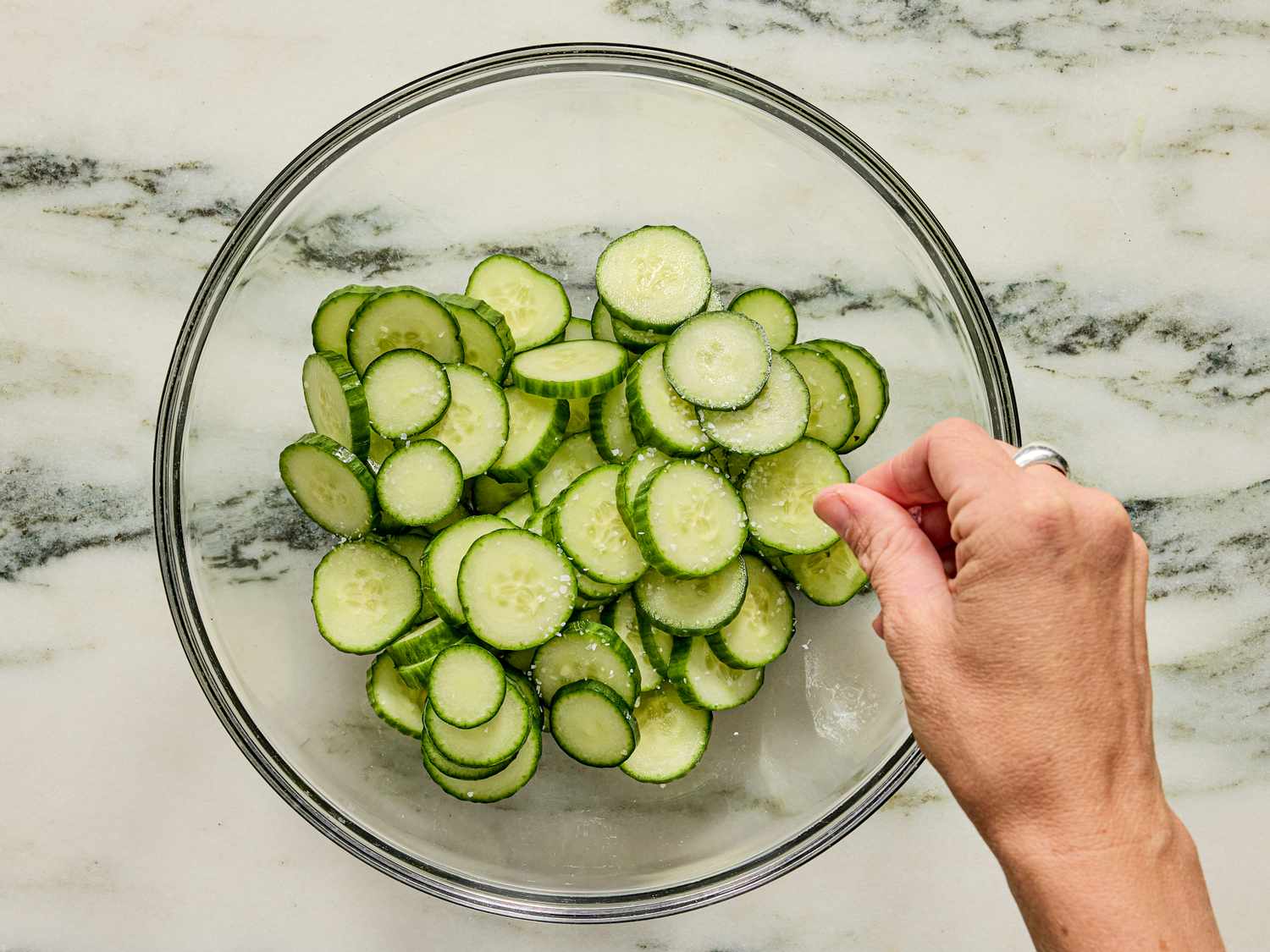 Hand adding seasoning to a bowl of sliced cucumbers