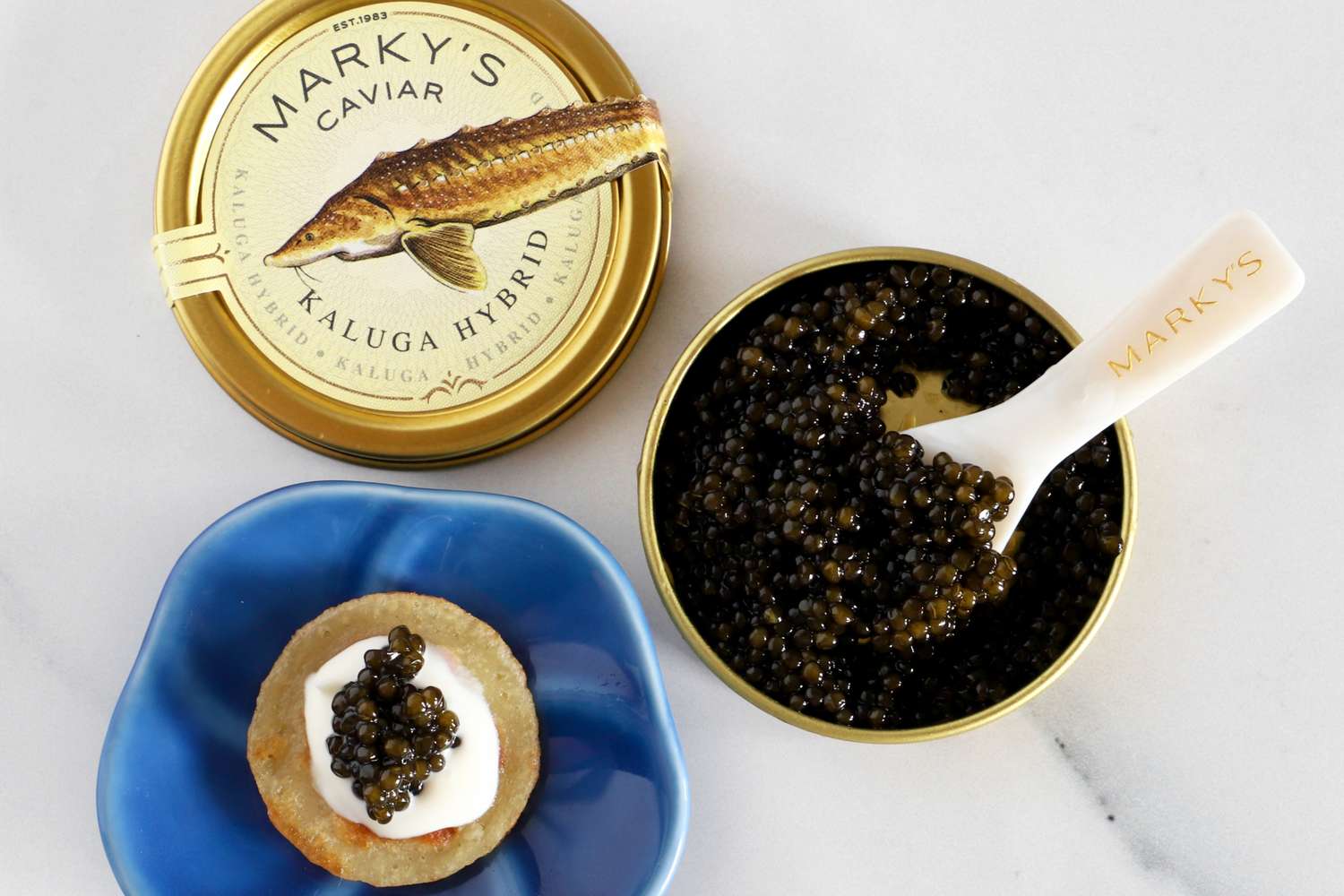 A tin of Marky's kaluga hybrid caviar next to a prepared blini topped with crème fraîche and caviar