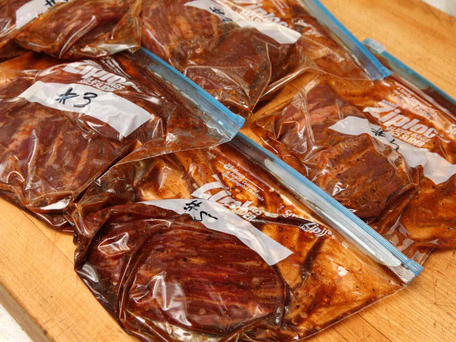 Labeled plastic zipper-lock bags of marinating skirt steak on a wooden cutting board.