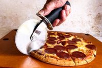 A hand holding an OXO pizza cutter while cutting a pepperoni pizza