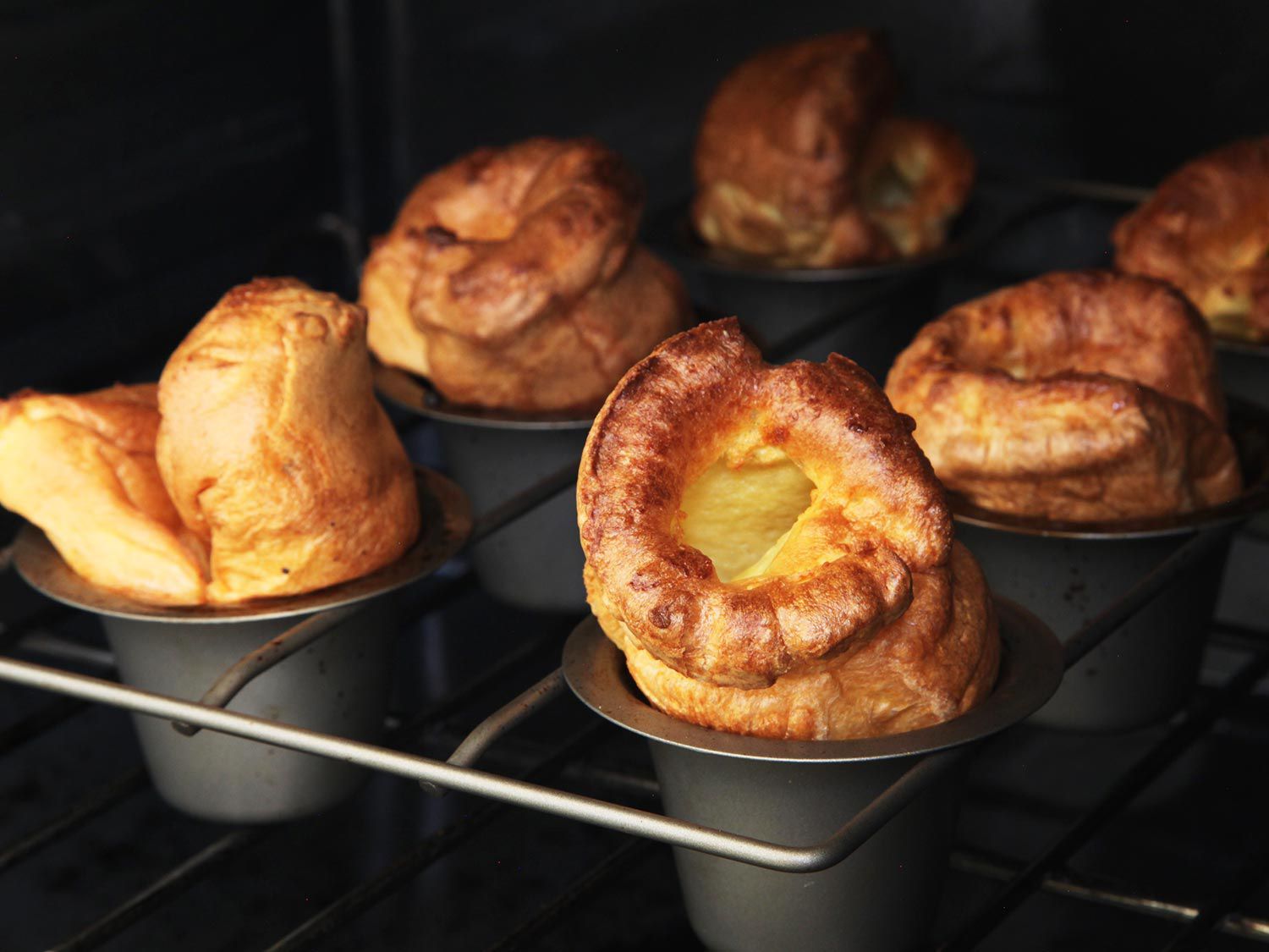 A pop over pan inside of an oven, with golden brown popovers emerging from the baking cups.