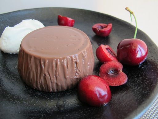 A chocolate blancmange unmolded on a plate, flanked by a mound of whip cream and fresh cherries.
