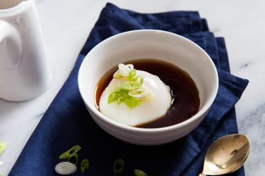 An onsen egg served at the table, cradled in a white bowl, half-submerged in soy-seasoned dashi. Sliced scallion is sprinkled over the top.