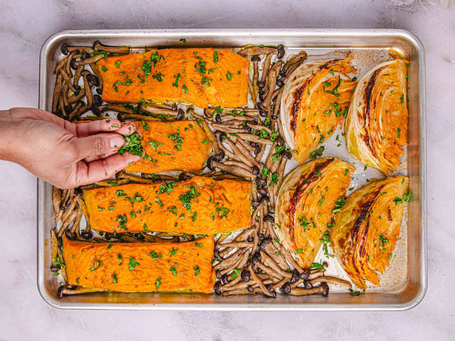 Sheet pan with salmon fillets, roasted mushrooms, and cabbage wedges being garnished