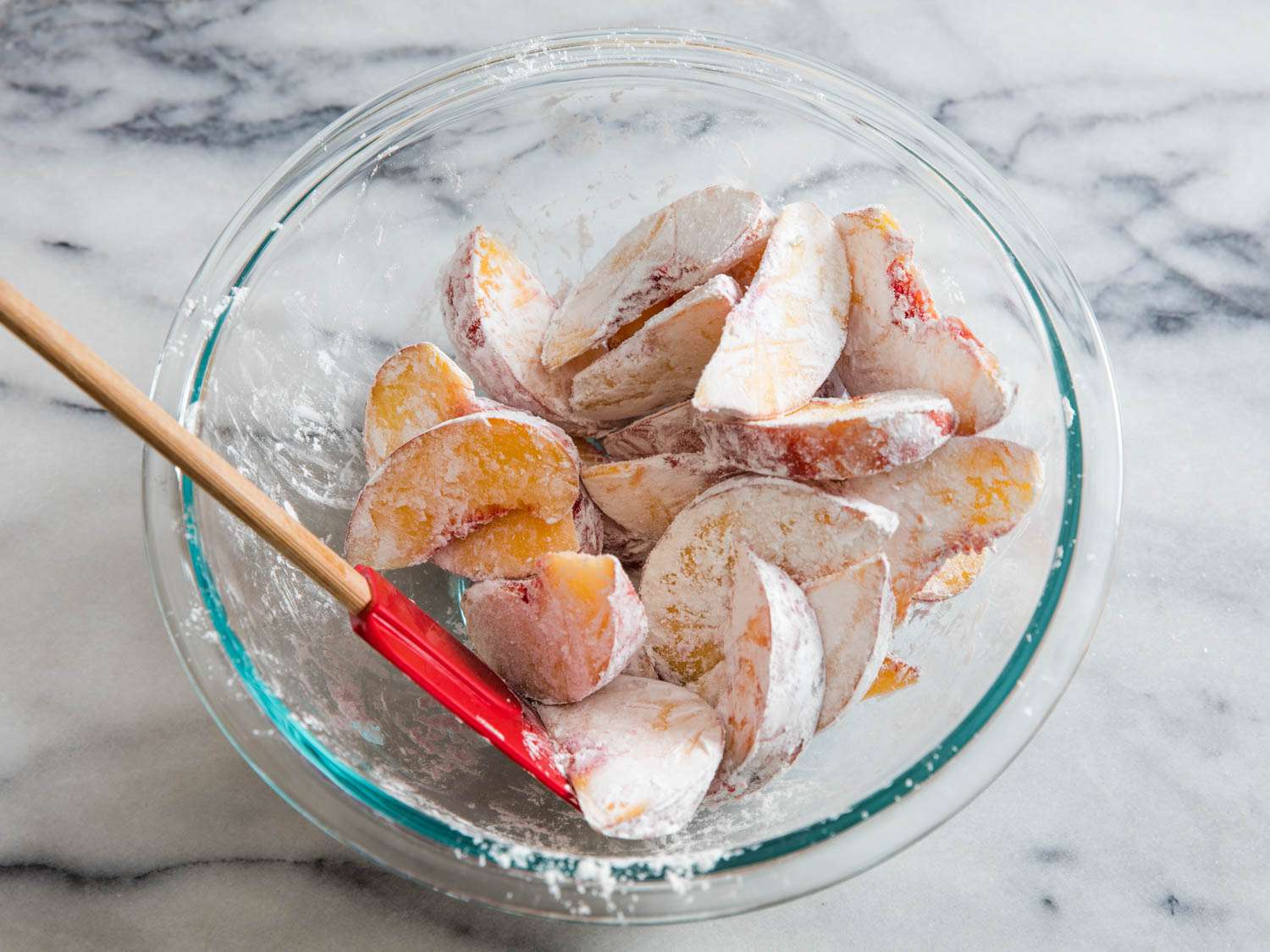 Peach slices are tossed with tapioca starch in a tmepred glass mixing bowl.