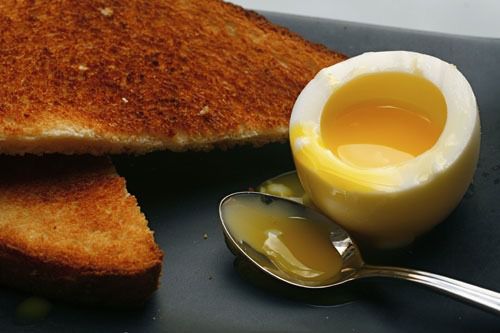 A soft-boiled egg cut open to reveal runny yolk next to a piece of toast and a spoon.