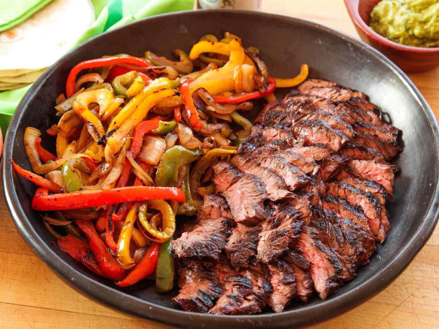 A bowl full of cooked peppers and onions and grilled sliced steak for fajitas, with tortillas and guacamole visible in the background.