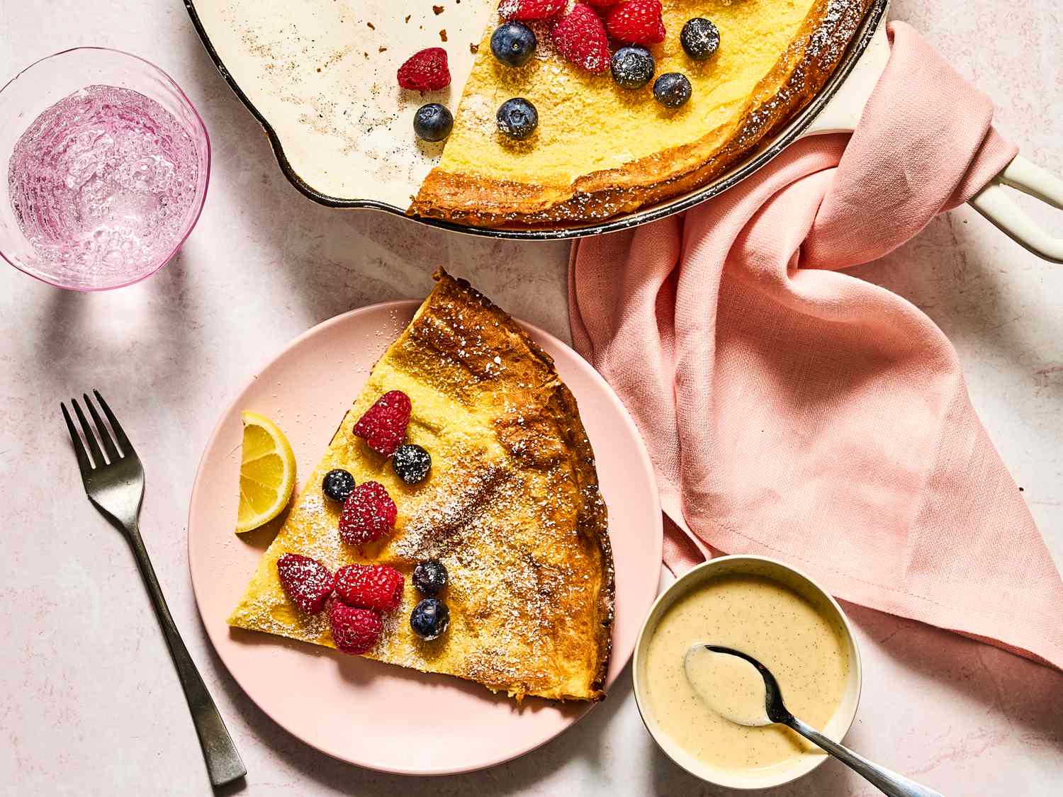 A Dutch baby pancake topped with mixed berries and powdered sugar on a plate and in a skillet, served with lemon wedge and sauce