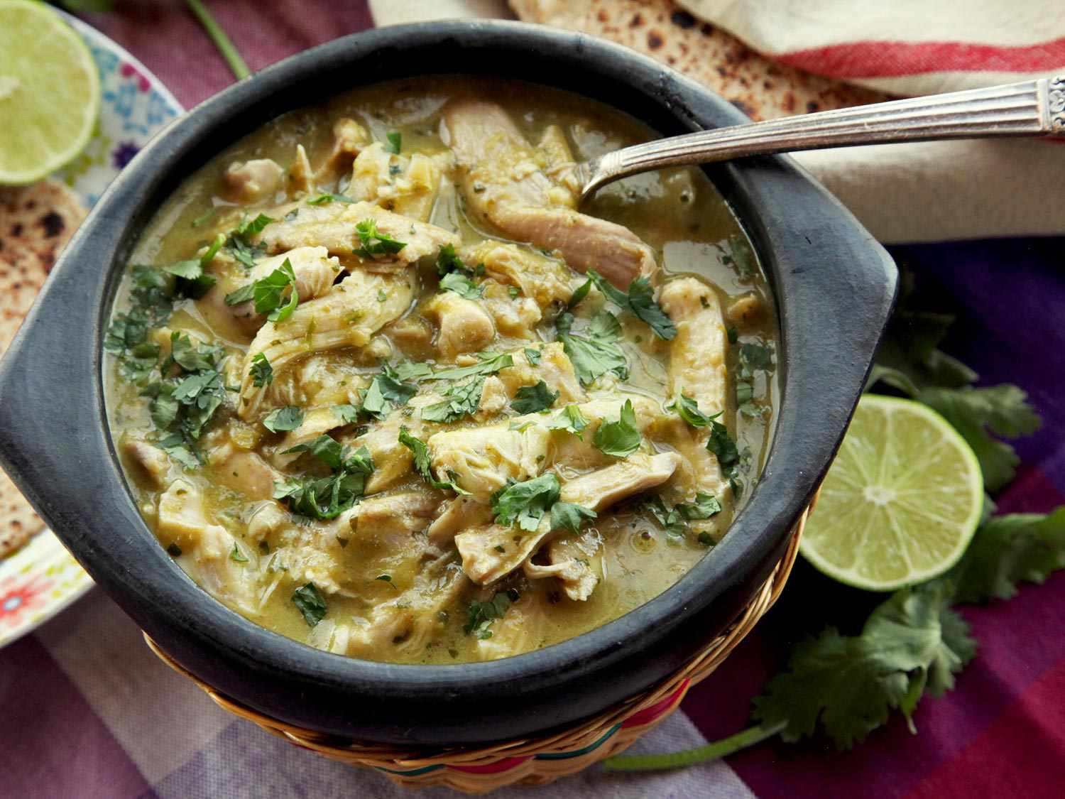 A bowl of chicken green chili (chile verde), garnished with cilantro, with tortillas and limes in the background.