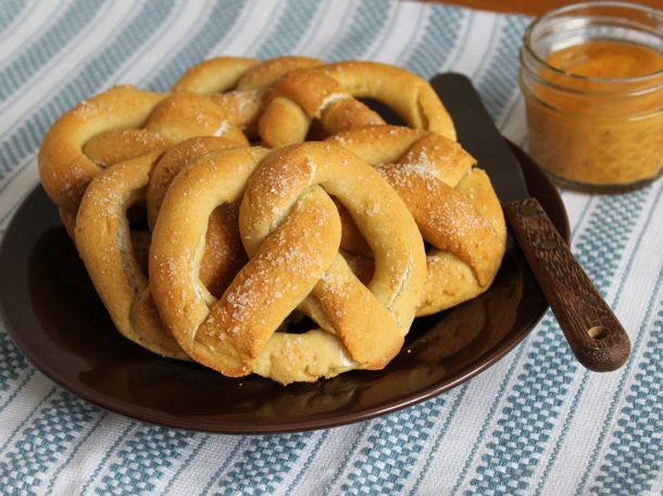 Three large gluten-free soft pretzels shingled on a plate, flanked by a spreader and a jar of mustard.