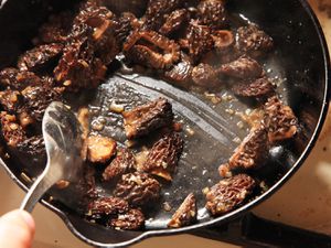 Sautéed morel mushrooms in a cast iron skillet being stirred with a large slotted spoon. 