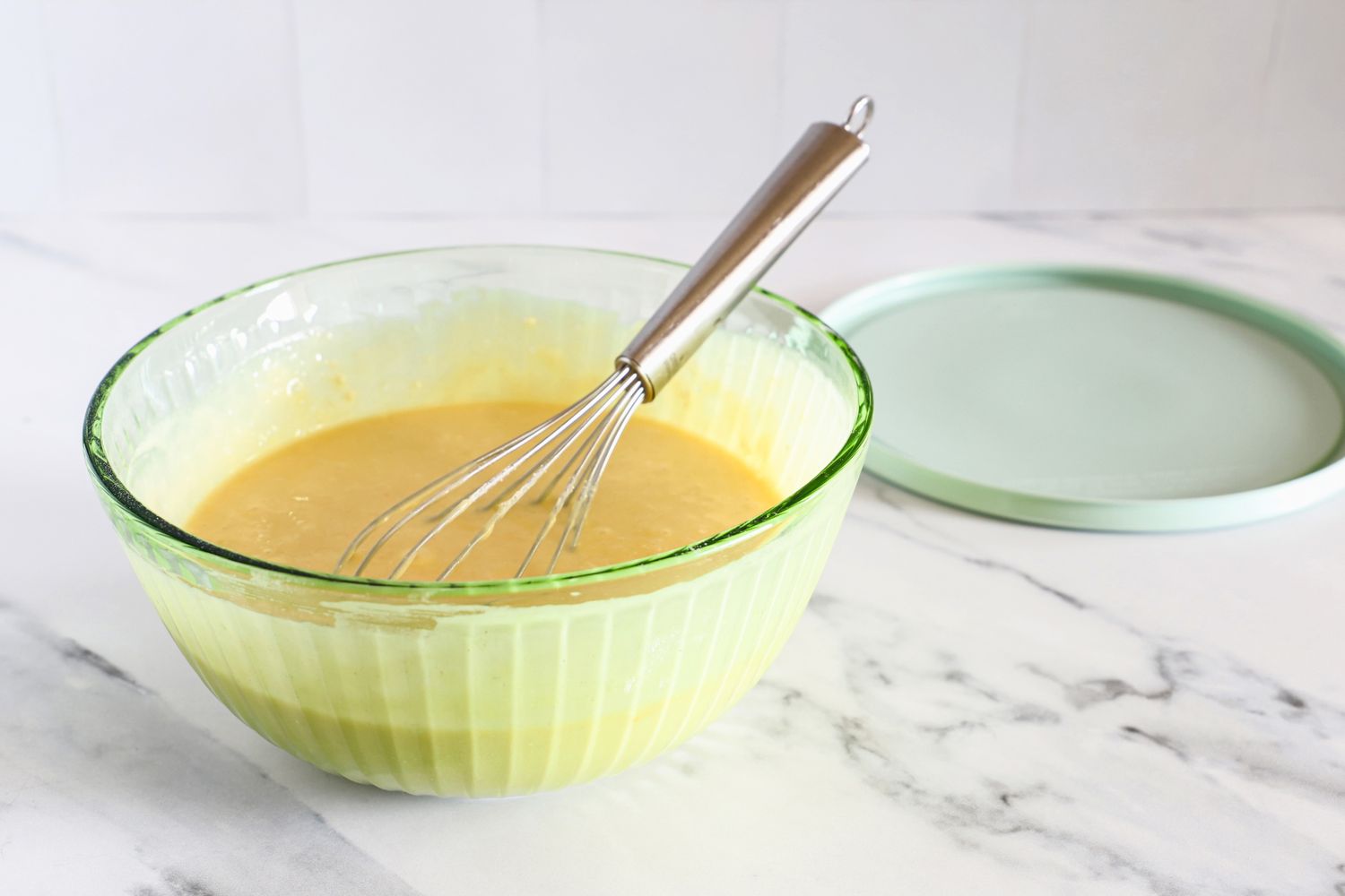 A Pyrex bowl containing batter with a whisk inside placed on a countertop next to a lid