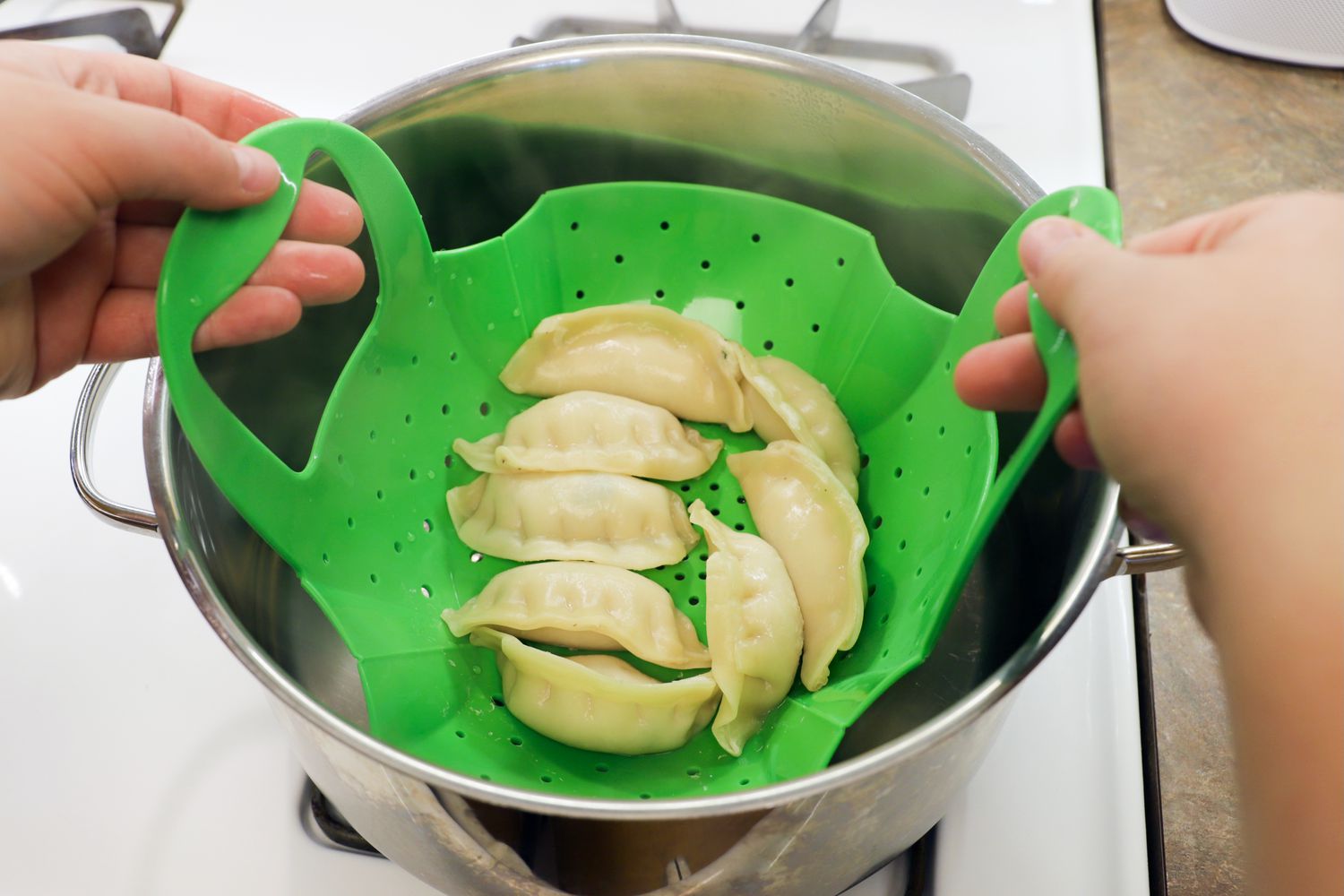 two hands holding the handles of a silicone steamer basket filled with dumplings and lifting it out of a pot
