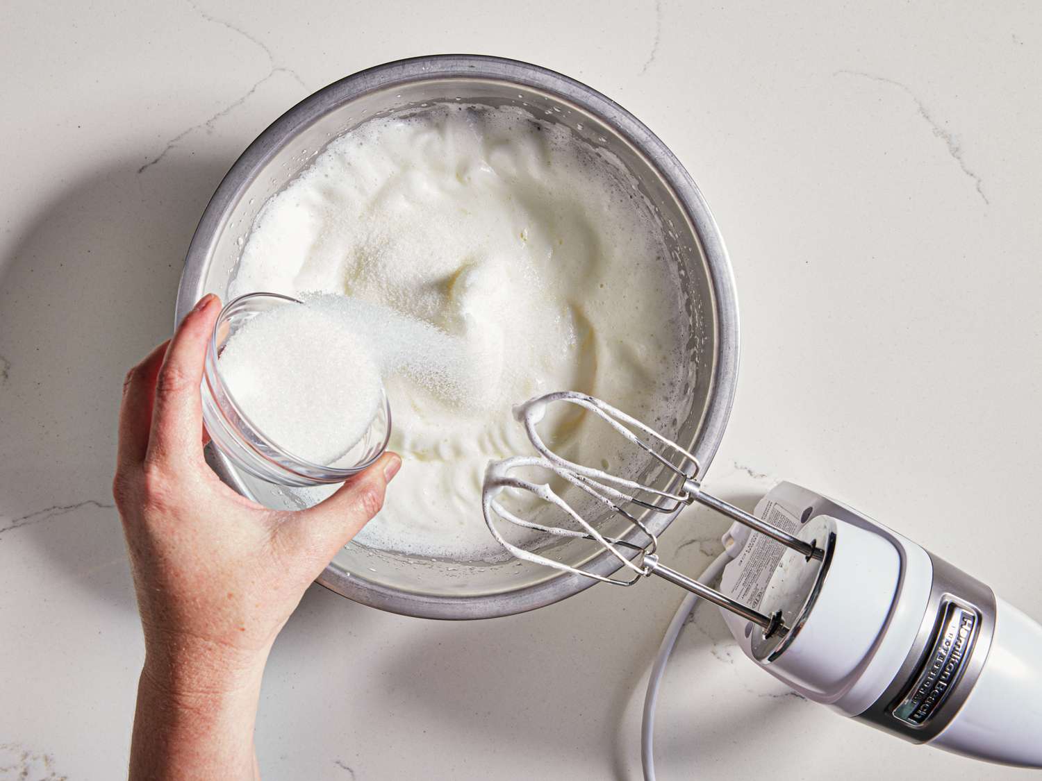 Sugar being adding to a bowl of whipped egg whites