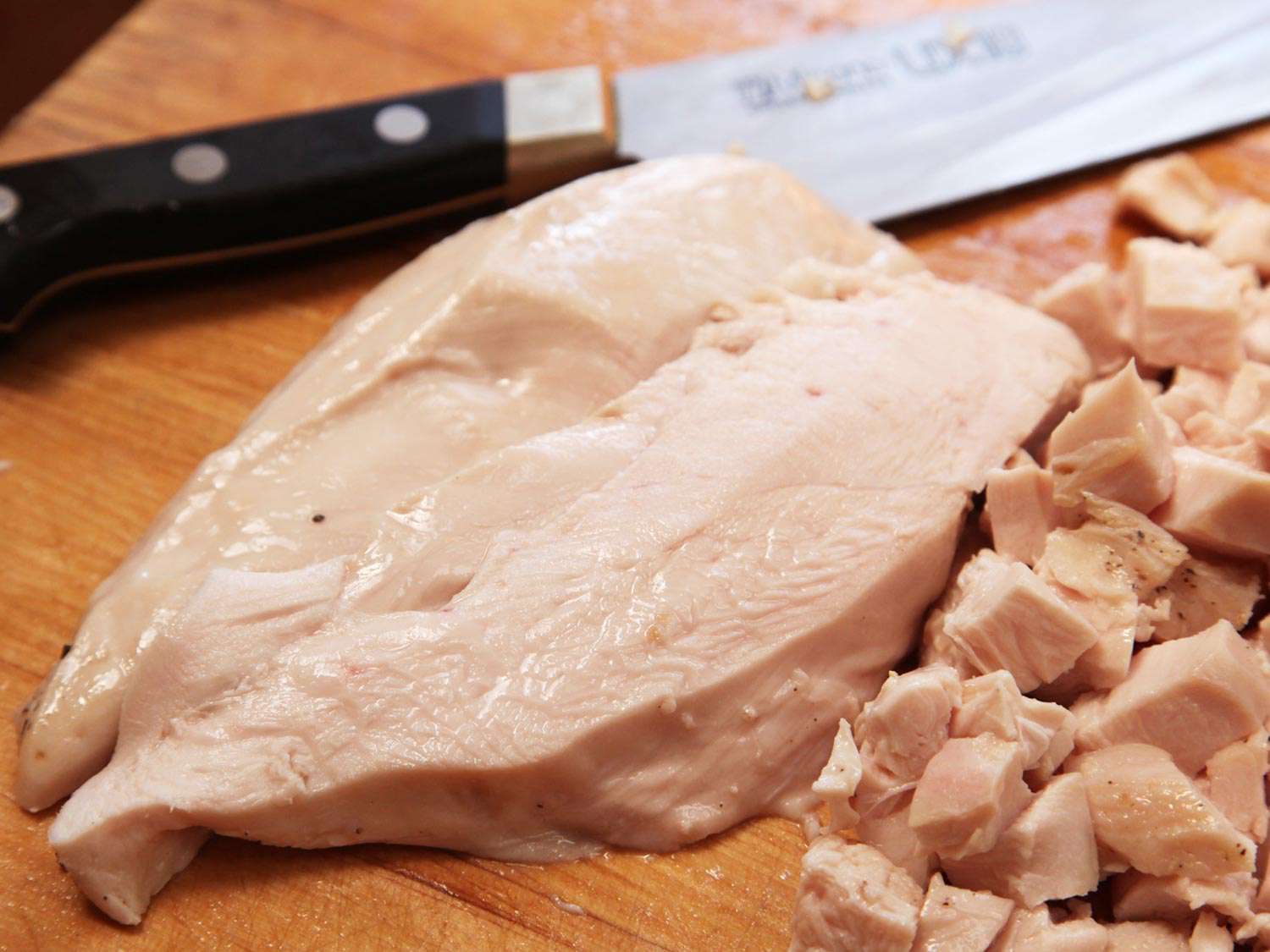 A whole chicken breast with the skin and bones removed, next to a knife and cubed, cooked chicken.
