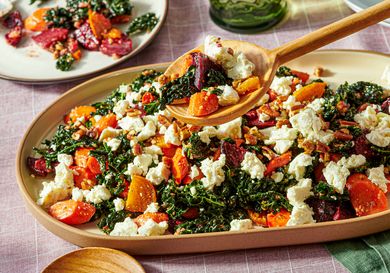 A platter containing kale salad with roasted root vegetables and crumbled cheese served with a wooden spoon
