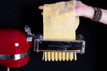 a hand feeding a sheet of pasta through a stand mixer's pasta cutter attachment