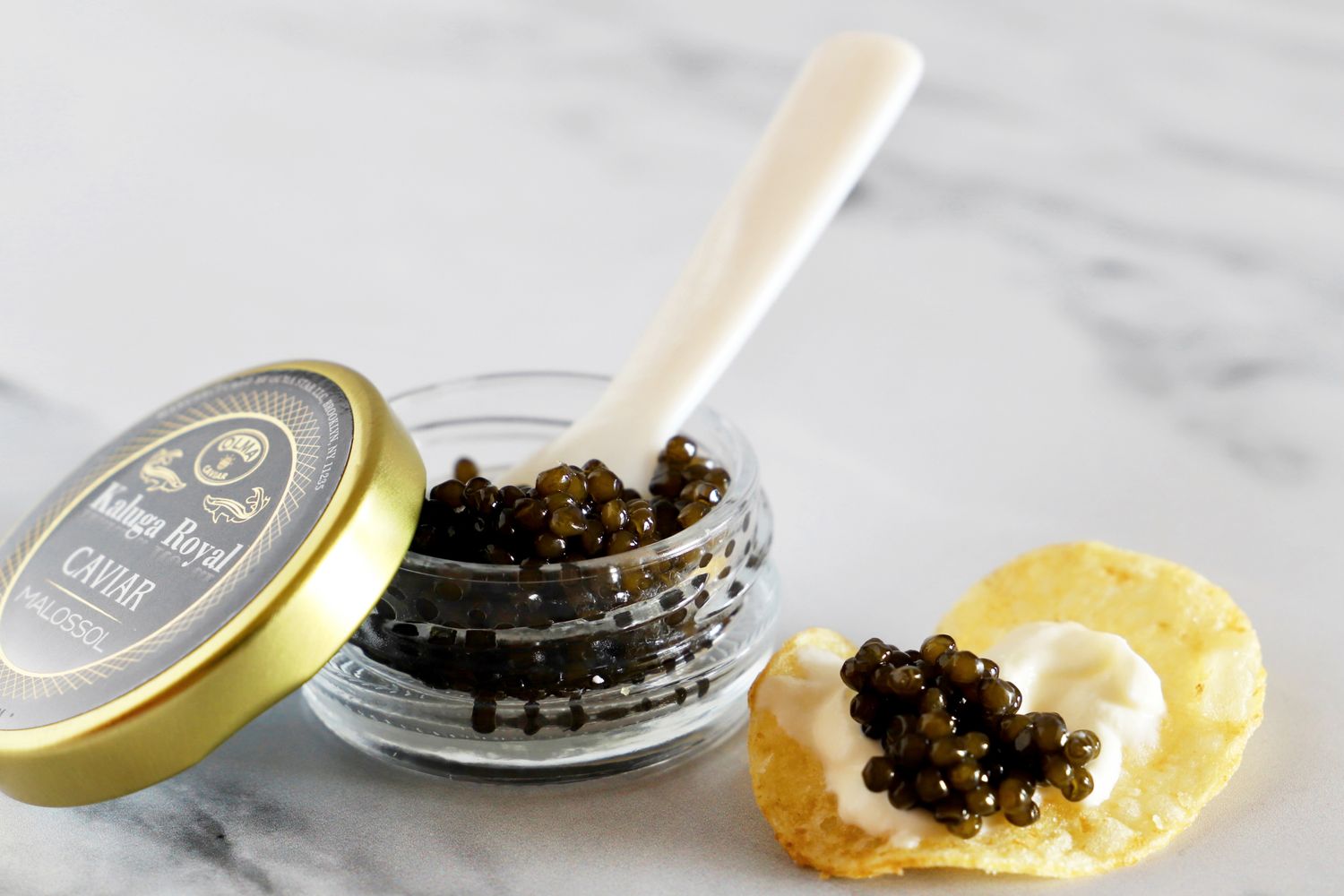 An open jar of Olma kaluga royal caviar next to a potato chip topped with crème fraîche and a scoop of caviar
