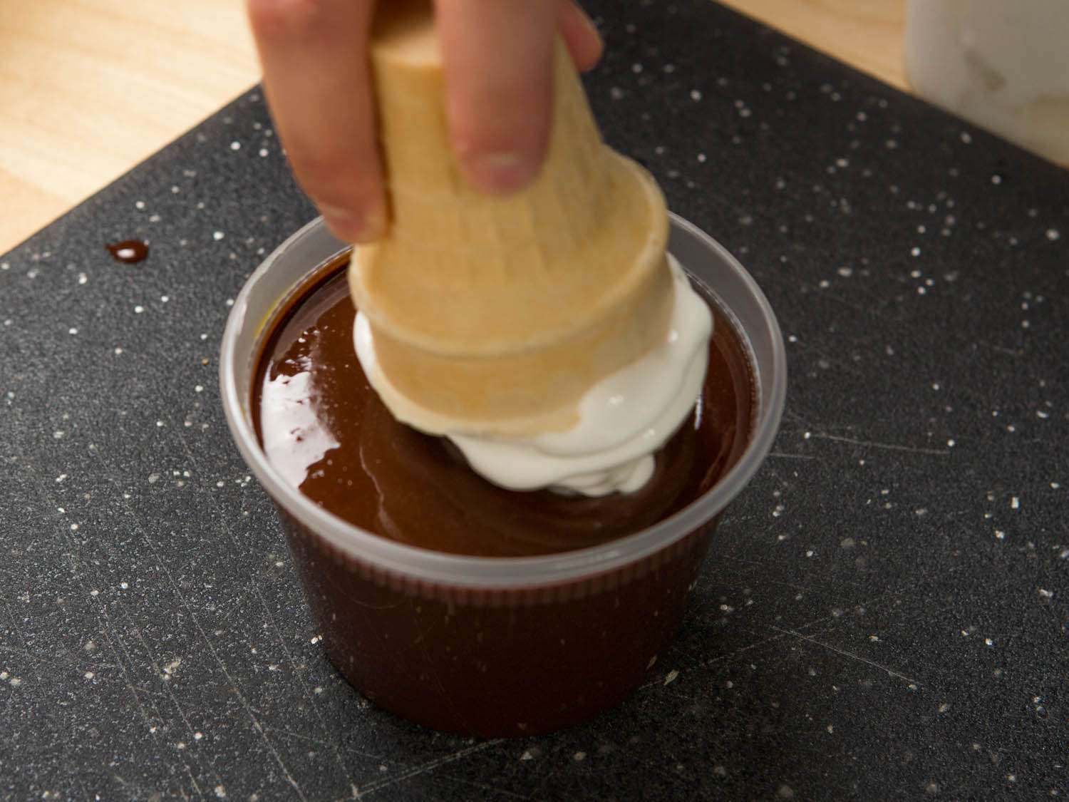 Hand dipping the top of a soft-serve ice cream cone into a plastic container of melted chocolate coating.