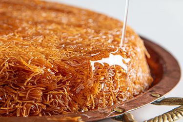 Knafeh being drizzled with syrup from above set on a round copper tray with handles